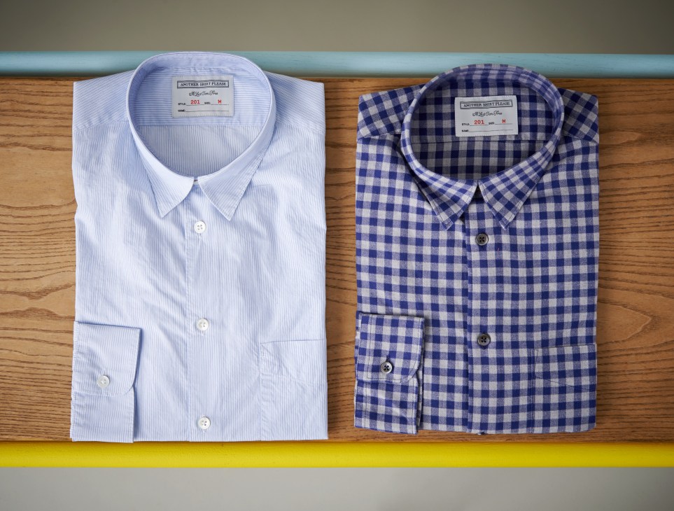Two folded shirts from Another Shirt Please displayed on wooden shelf: light blue striped and navy gingham check.