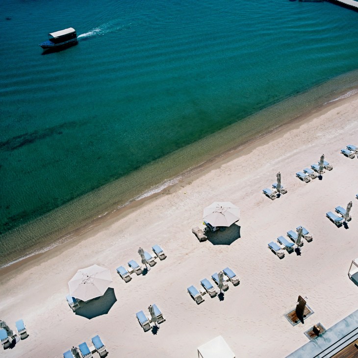 Aerial view of a pristine beach with organized rows of blue and white lounge chairs, white umbrellas, and a motorboat in turquoise waters.