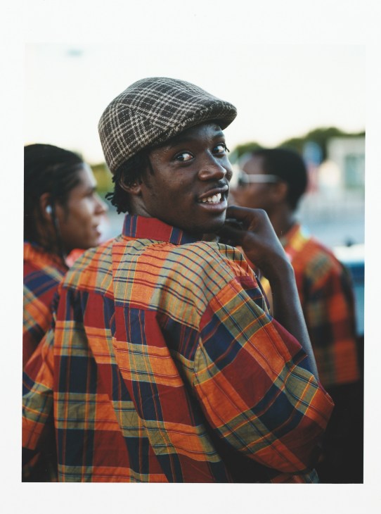 A smiling Mozambique athlete wearing a red and yellow plaid shirt and patterned cap at the opening ceremony.