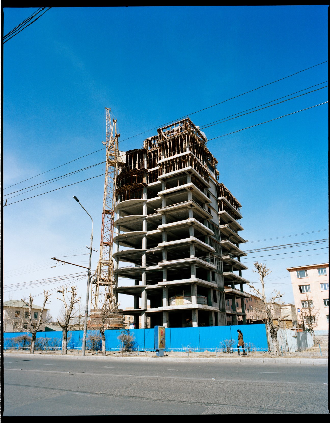 Half-finished concrete residential tower under construction in Ulan Bator with blue hoarding and crane visible.