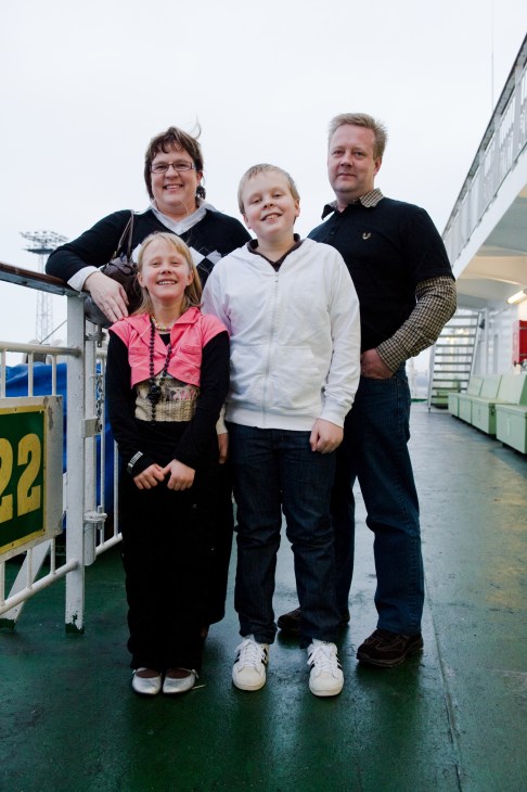 A family of four stands on the deck of a ferry, smiling at the camera with the ship's white superstructure visible behind them.