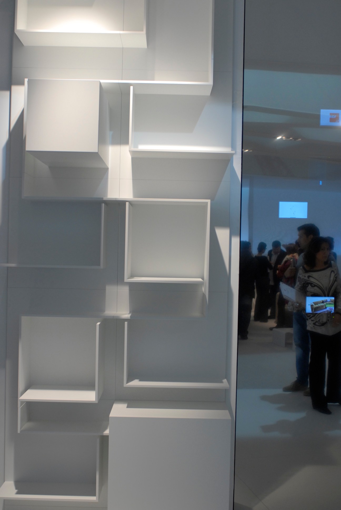 White modular shelving display with illuminated compartments at a design fair, visitors viewing in background.