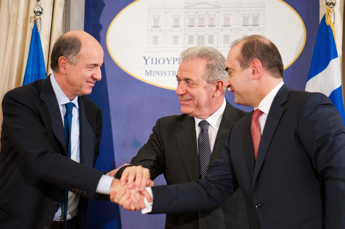 Three men in business suits shake hands at a government ministry event with flags in background.