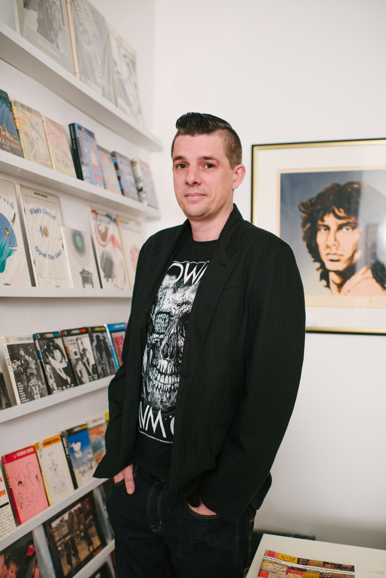 Josh Carey stands in his bookshop surrounded by shelves displaying vintage books and framed artwork.