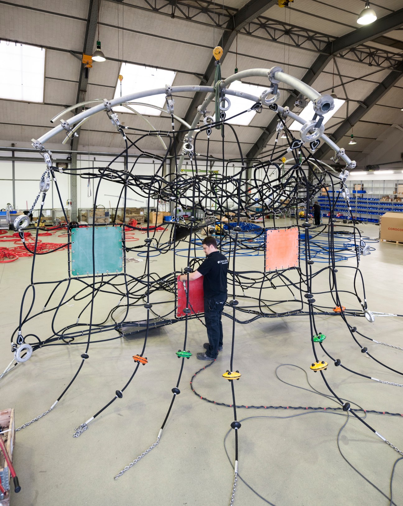 Worker assembling a geometric dome-shaped Corocord playground net structure in factory with metal frame and black ropes.