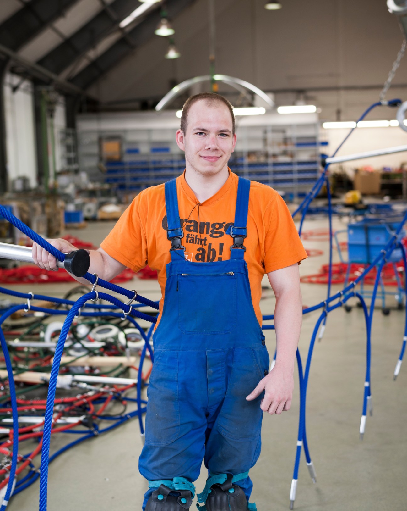 Benedikt Klauss, a young assembly team assistant wearing blue overalls and orange shirt, holds a blue rope in Corocord's Berlin factory.