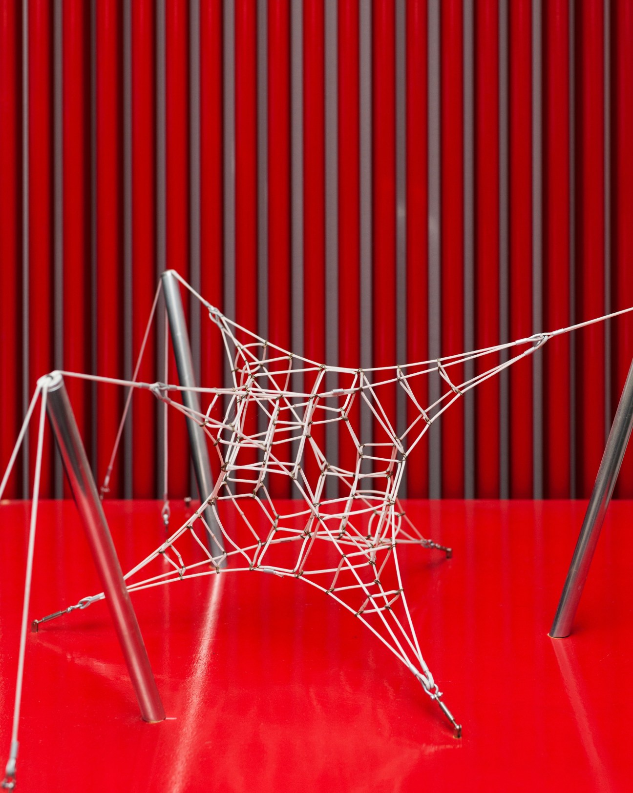 Miniature white rope net model with geometric structure on red surface against red corrugated backdrop.
