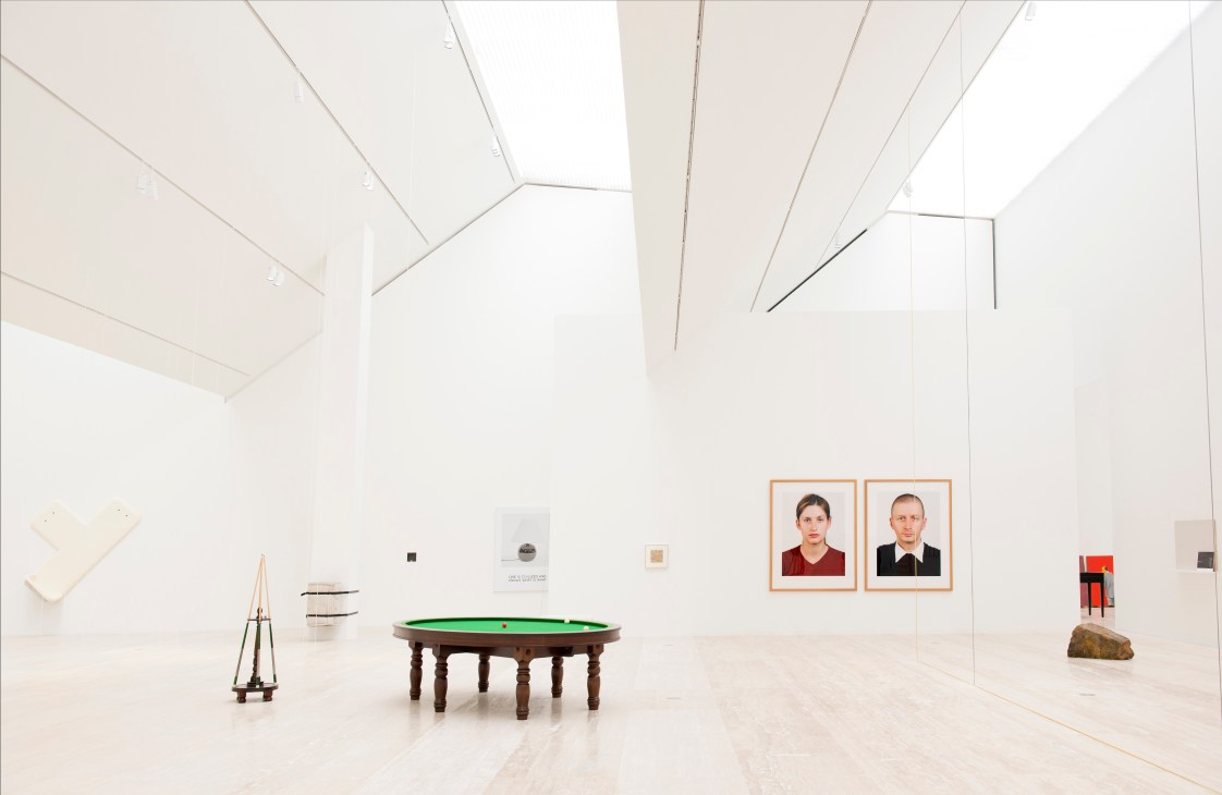 A bright gallery space with skylights displaying contemporary artworks including a green billiard table and framed portraits.