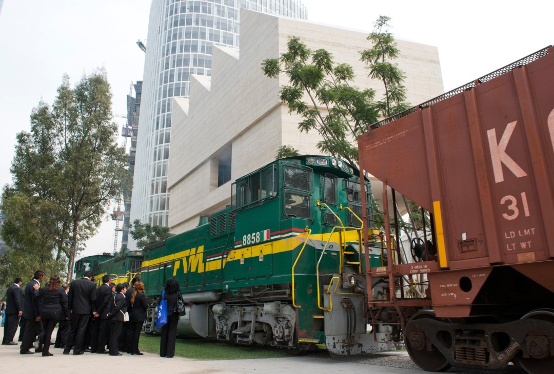 A green and yellow freight train next to a red boxcar with formally dressed visitors standing nearby in front of modern buildings.