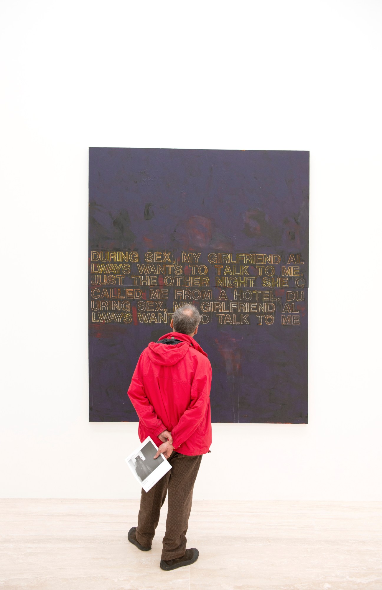 Visitor in red jacket viewing a Richard Prince artwork with text on dark background.