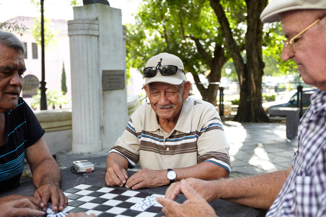 Three men playing checkers in a shaded San Juan park on a sunny day.