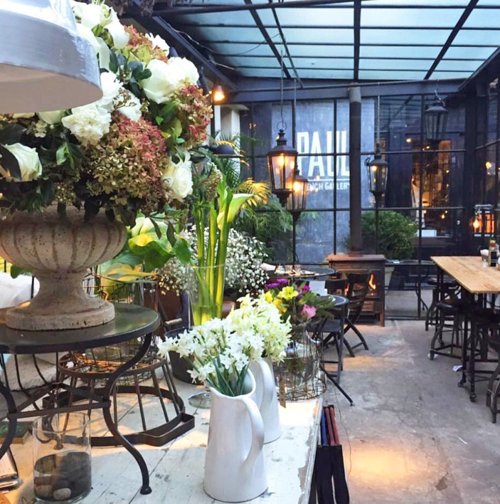 Paul French Gallery's open-air corridor with flowering planters, bistro seating, and glass ceiling.