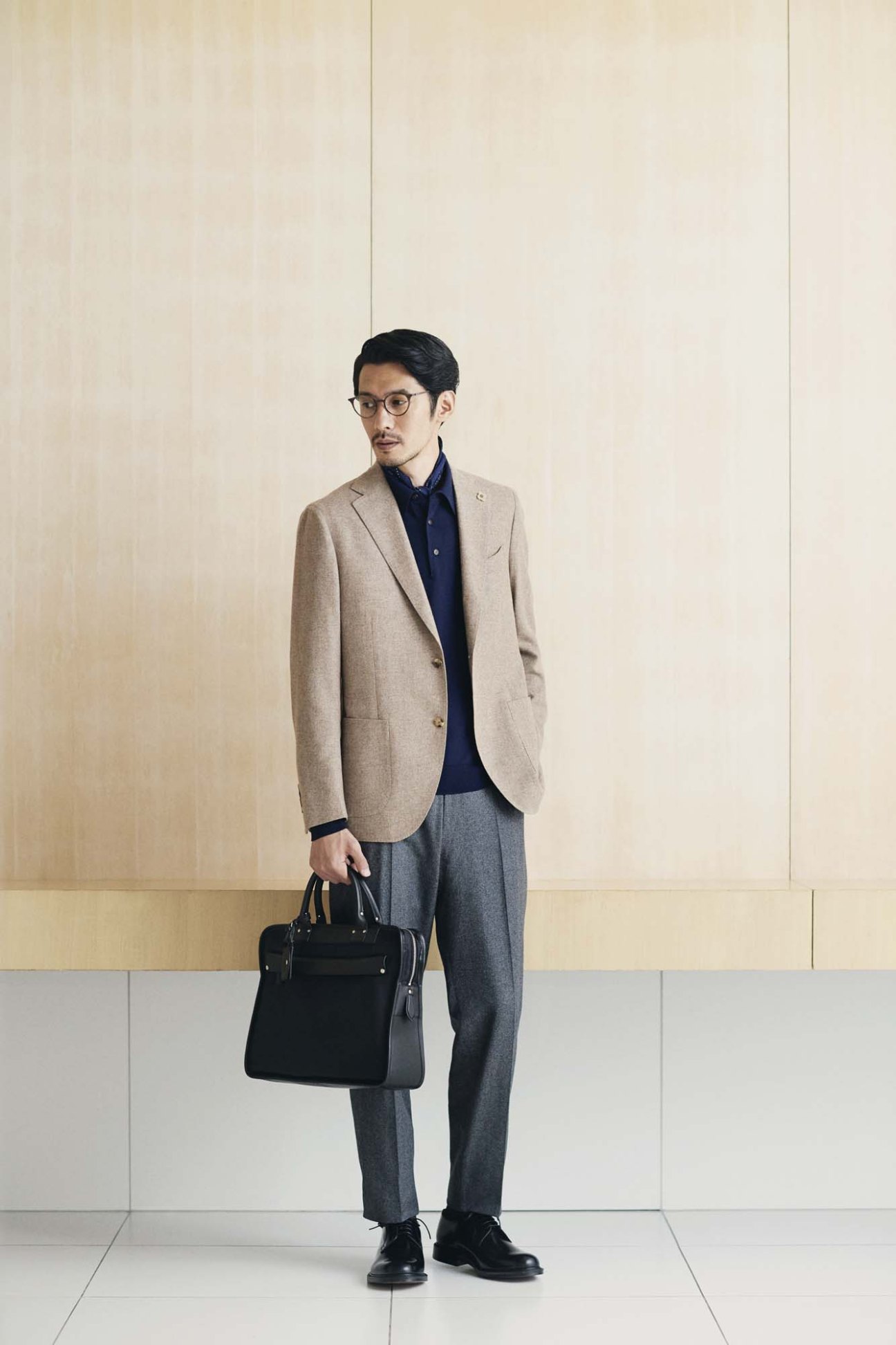Man wearing beige blazer, navy polo shirt, grey trousers, glasses and holding black leather briefcase.