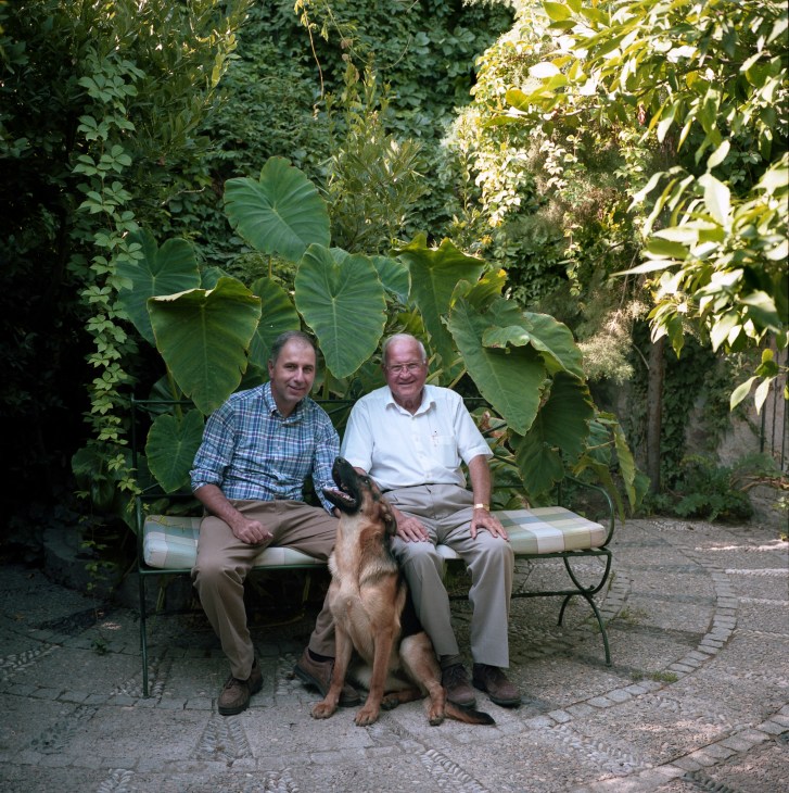 Reggie and Antony Gallia sit on a bench in a lush garden with a dog between them.