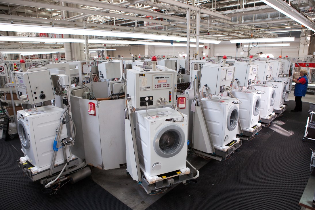 Miele washing machines and dryers lined up in a factory facility during quality testing with workers in the background.