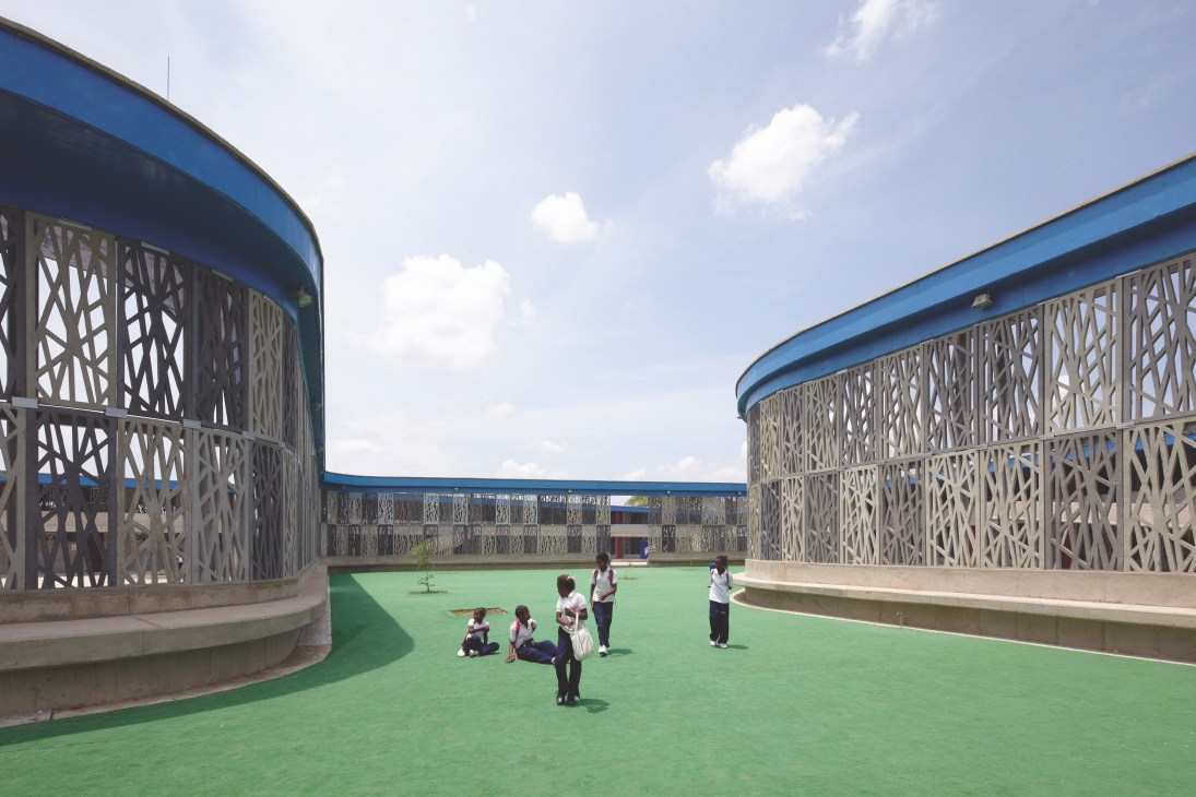 Children playing in a curved outdoor playground surrounded by decorative concrete fretwork walls under blue sky.