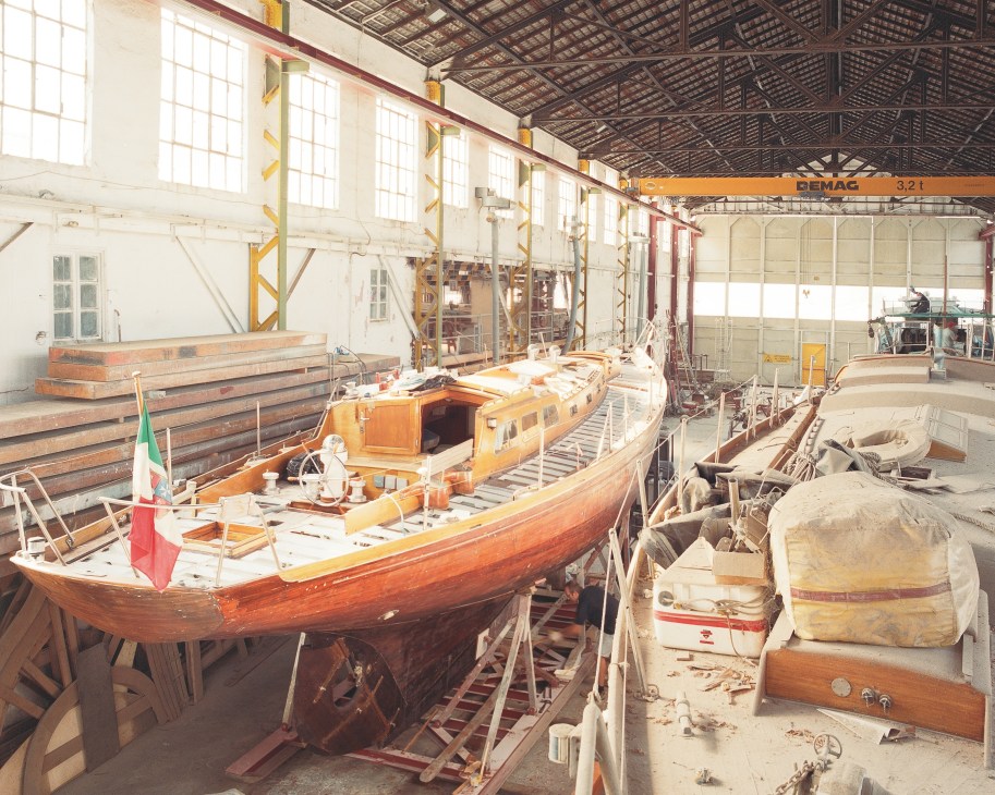 Wooden sailing yachts under restoration in a boatyard workshop with industrial ceiling and natural lighting.