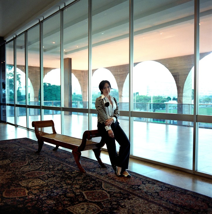 Aletha Pennati Migita stands in a modern corridor with floor-to-ceiling windows overlooking Brasília's landscape with distinctive architectural arches visible through the glass.