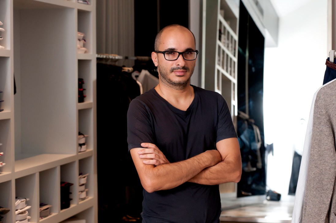 Sketch designer Yossi Katzav stands with arms crossed in his boutique, wearing glasses and a black v-neck shirt.