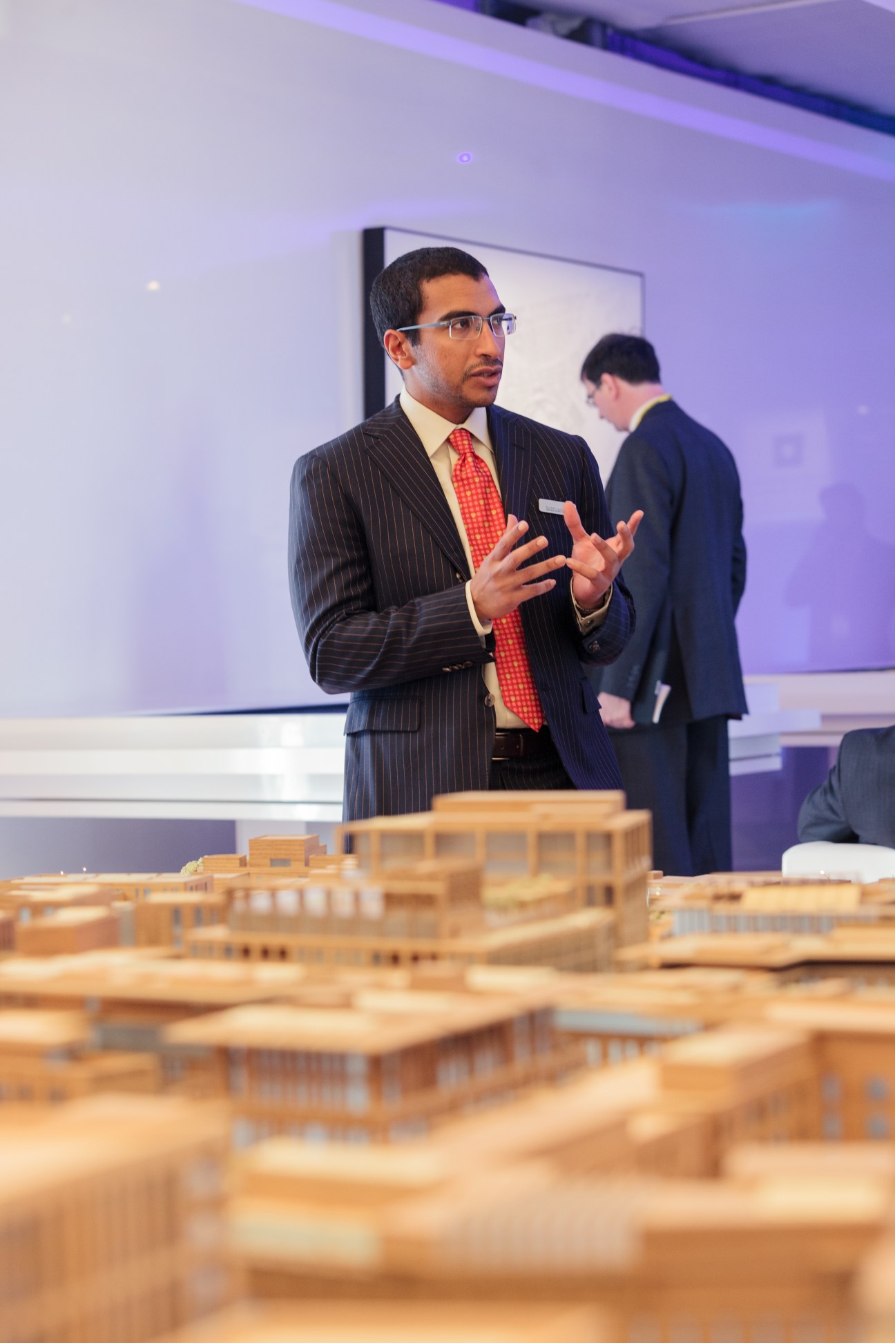 Man in pinstriped suit and red tie gestures while presenting beside a wooden architectural model at Qatar's Mipim pavilion.
