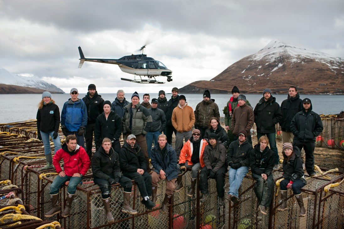 Production crew from 'Deadliest Catch' TV show posing on crab pots in Unalaska with a helicopter and snow-capped volcano in background.