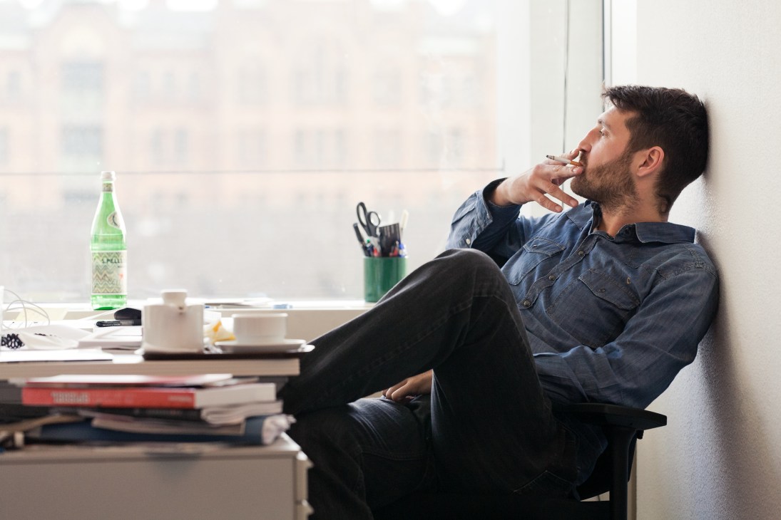Man in denim shirt sits at desk with feet up, smoking cigarette near window with city view.