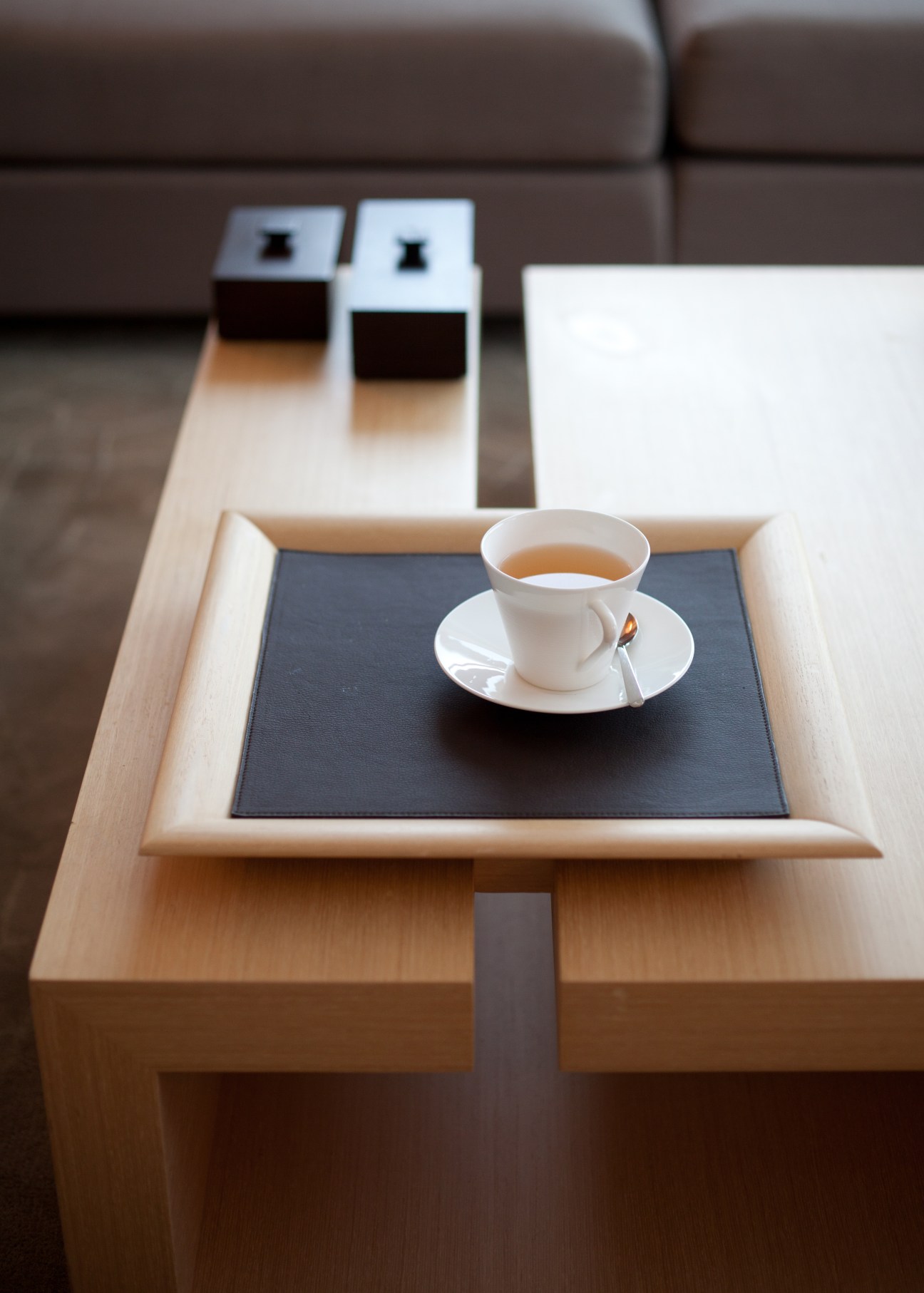 White porcelain teacup and saucer with tea on a black tray within a light wood frame, positioned on a wooden table with brown sofa in background