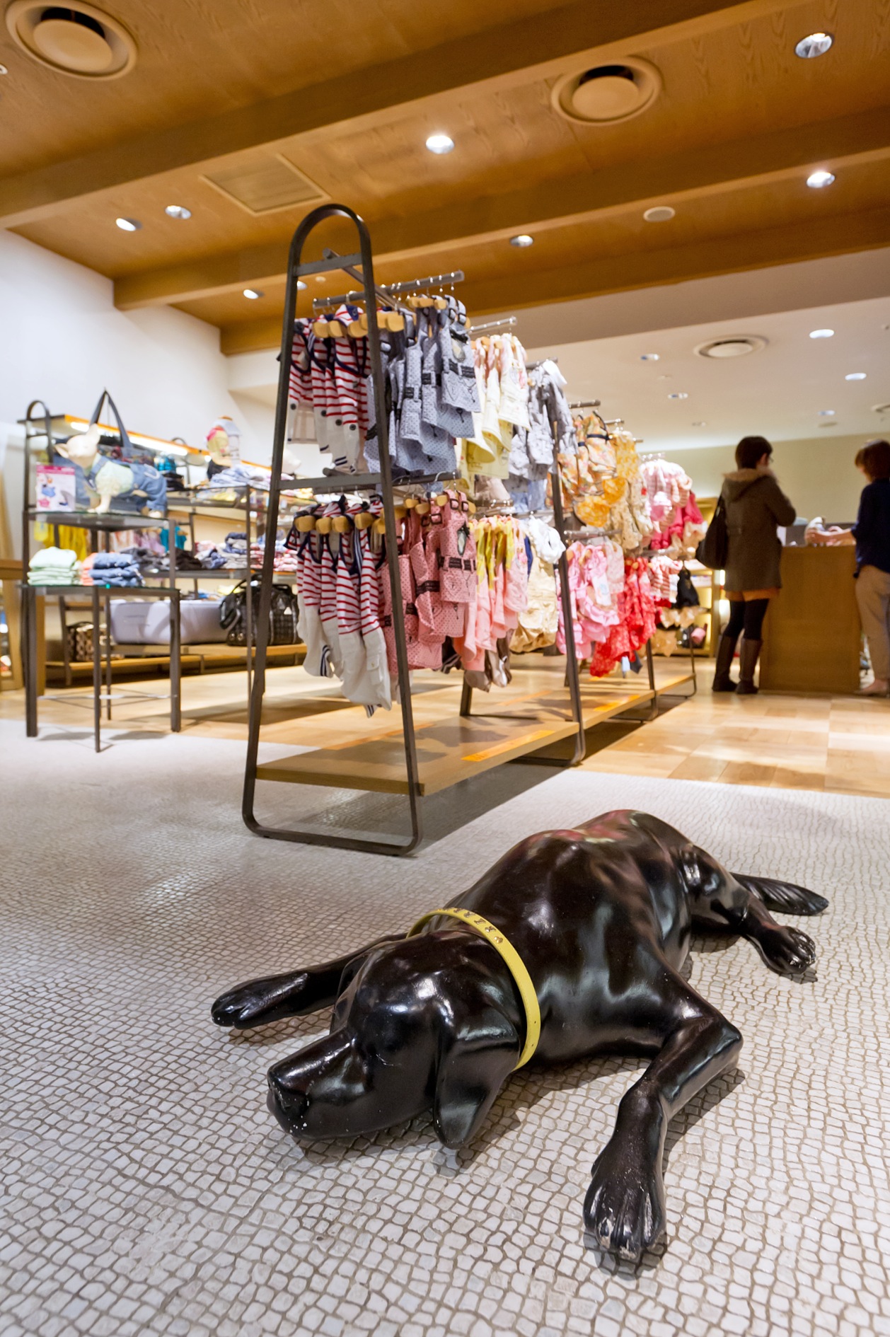 Black dog sculpture lying on floor in Hannari shop with colorful dog clothing displayed on racks behind it.