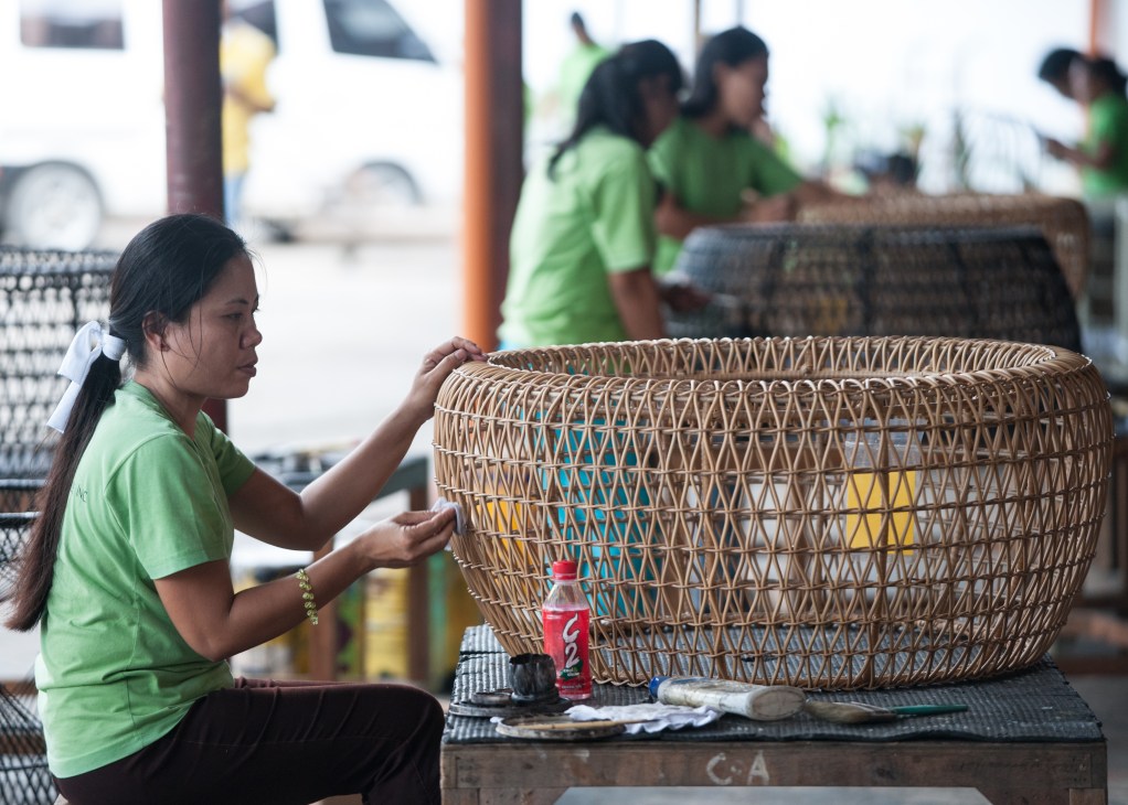 Woman in green shirt weaving a large rattan basket at a furniture factory in Cebu.