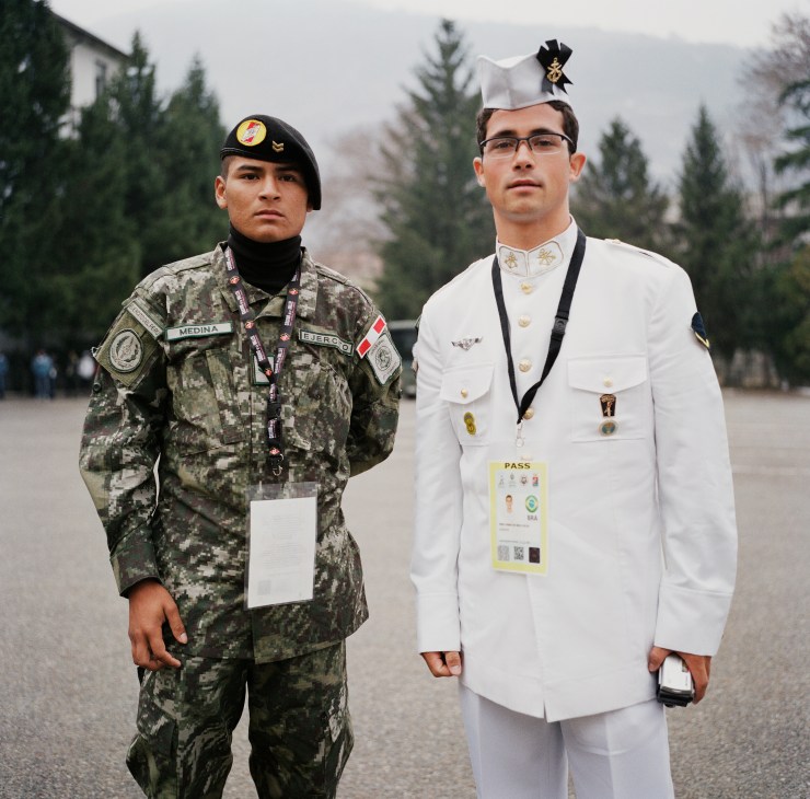 A military officer in camouflage uniform stands next to a naval officer in white dress uniform, both wearing credential passes.