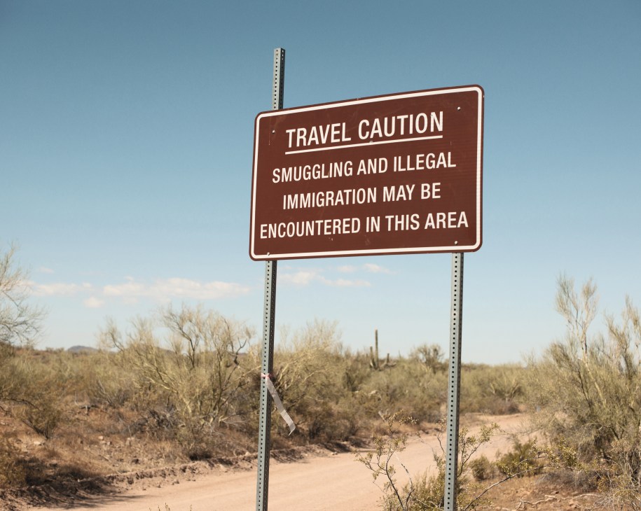 Brown warning sign reading 'Travel Caution: Smuggling and Illegal Immigration May Be Encountered in This Area' in Arizona desert landscape.
