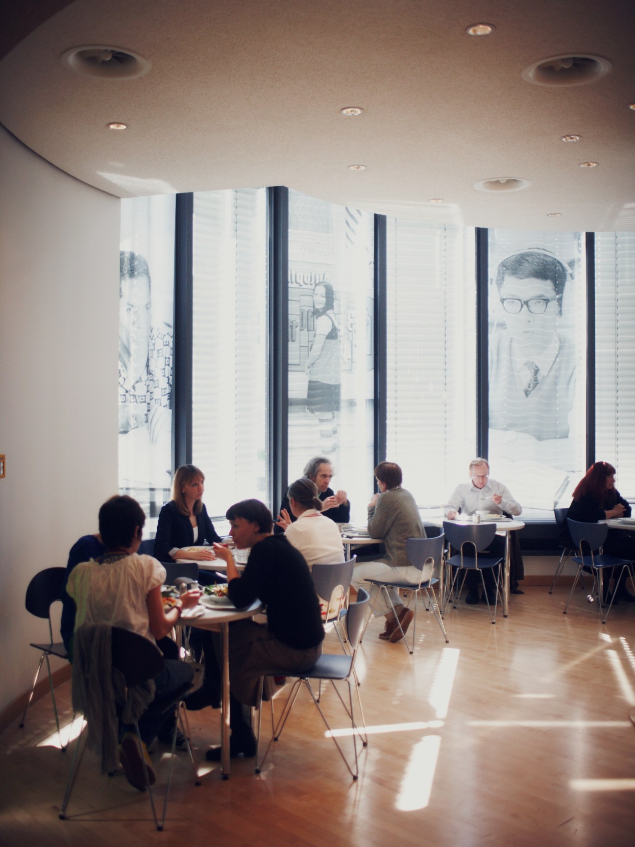 People seated at tables in a bright, modern café with large windows and portrait photographs on the walls.