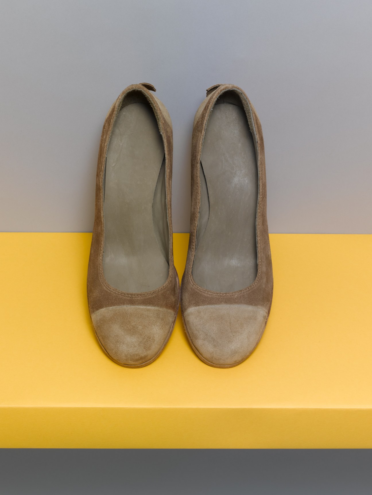 Pair of taupe suede women's shoes with two-tone coloring displayed on a yellow platform.
