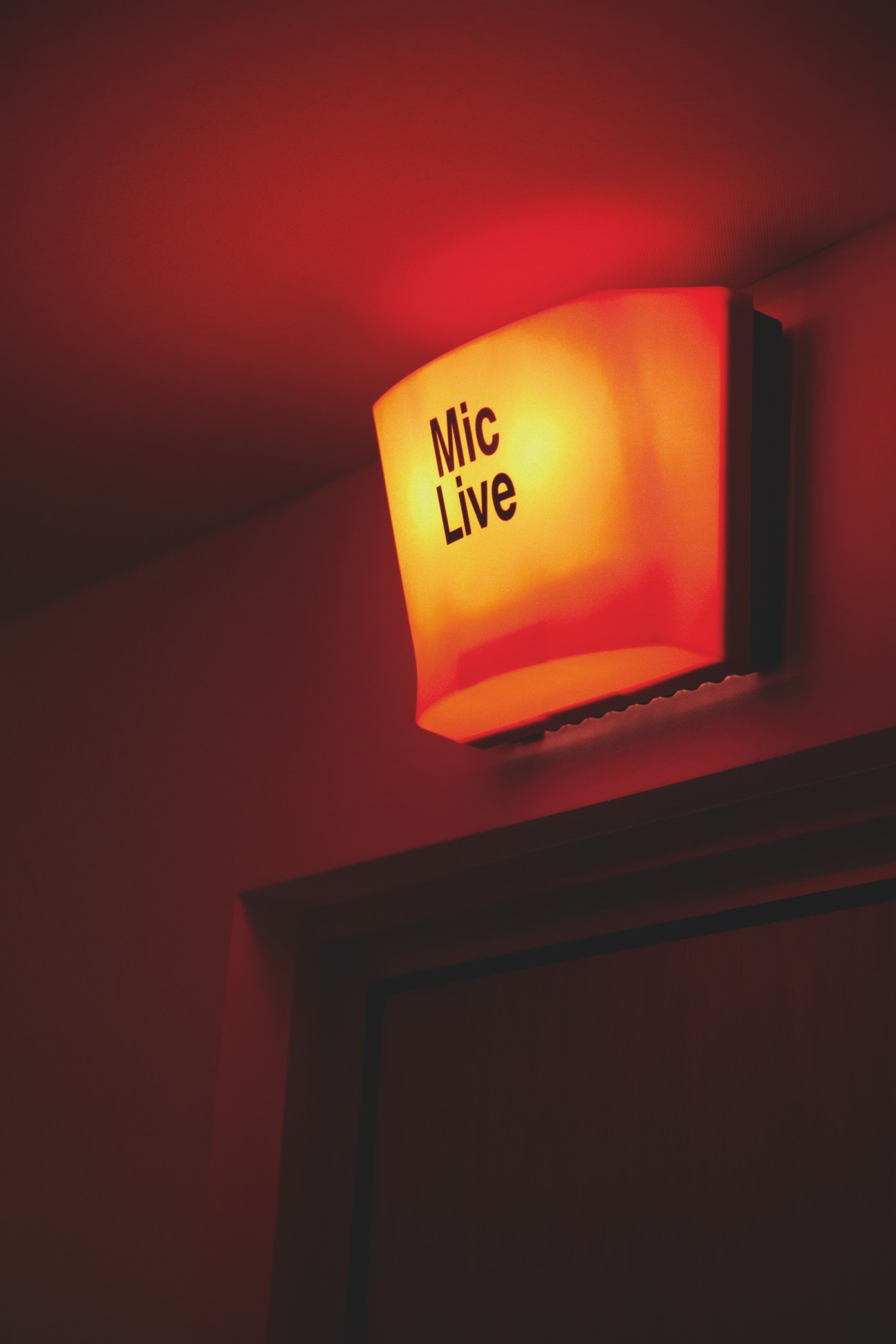 Illuminated 'Mic Live' sign glowing yellow and red against a dark red wall in Monocle 24 studio.