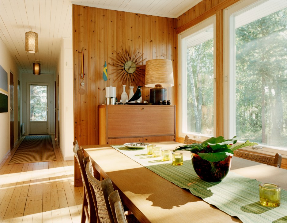 Wooden dining table and chairs in sunlit room with wood-paneled walls and large windows overlooking forest.