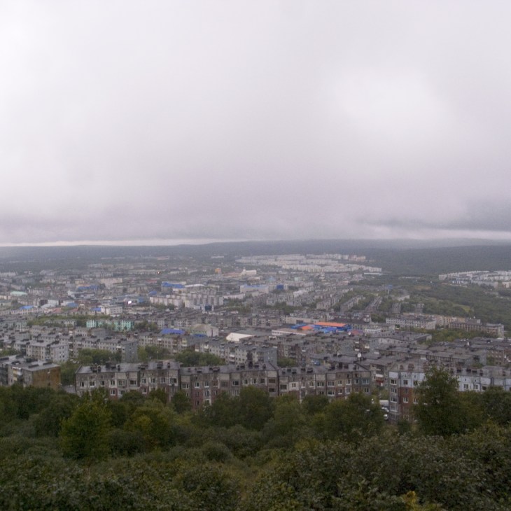 Aerial view of Petropavlovsk-Kamchatsky under overcast skies with Soviet-era apartment buildings and bay visible in the distance.