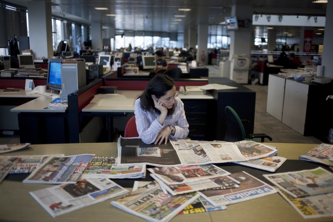 Woman reviewing Turkish newspapers at desk in modern media office with computers in background.