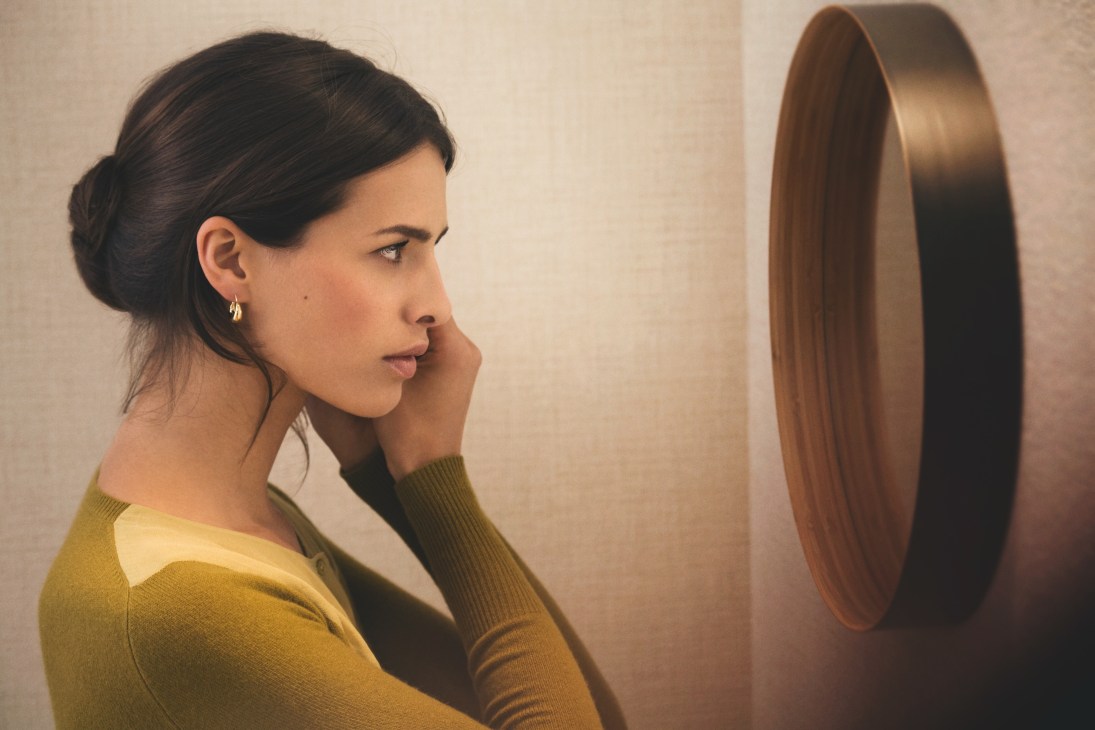 Woman in profile wearing a yellow cardigan and Georg Jensen earrings, looking at a circular mirror.
