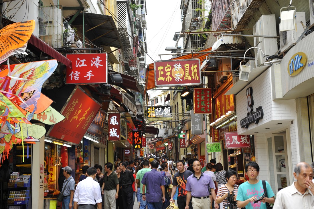 Crowded pedestrian street in Macau with Chinese shop signs, colorful lanterns, and traditional storefronts.