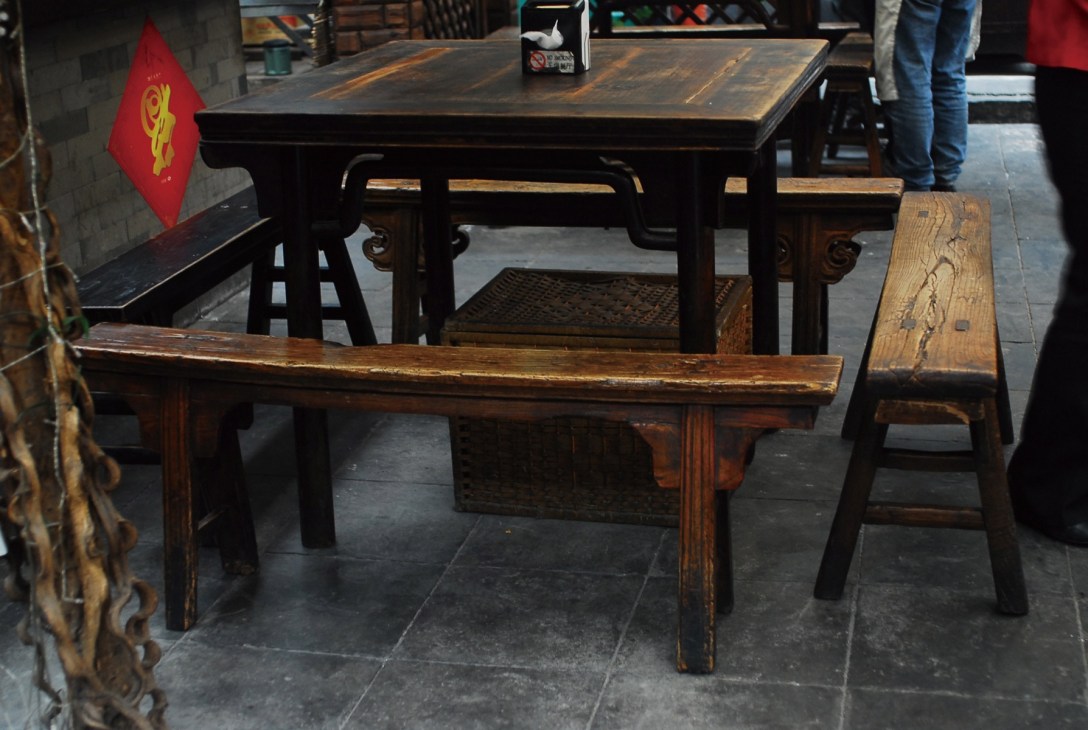 Traditional dark wooden benches displayed at an outdoor market or showroom with a red flag visible in background.