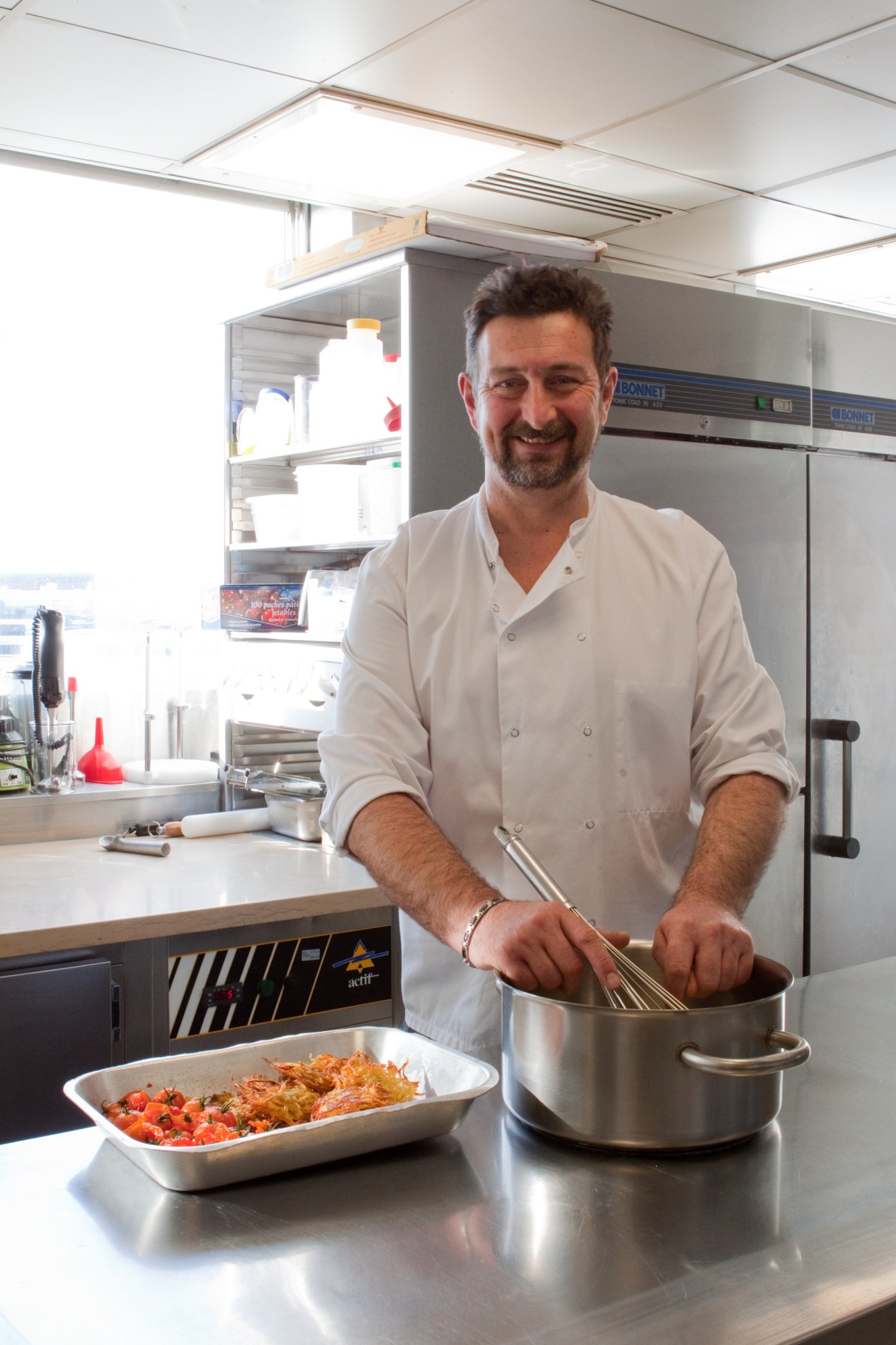 Chef Emmanuel Lejeune in a kitchen wearing a white chef's coat, holding a whisk over a pot with a prepared dish nearby.