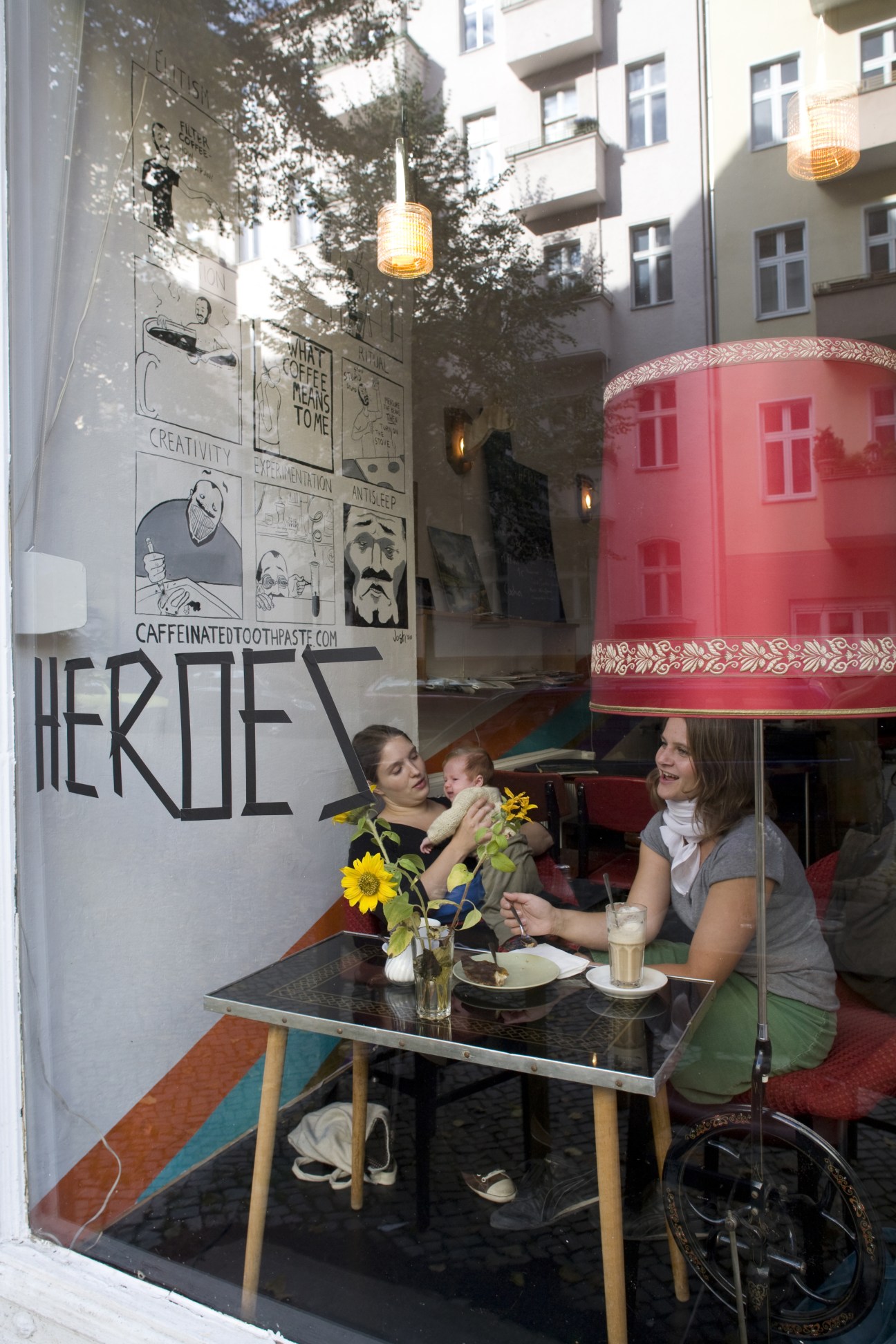 Two women and a baby sit at an outdoor table at Heroes café in Neukölln, with sunflowers and drinks on the table.