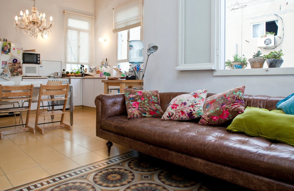 Bright living room with brown leather sofa, floral cushions, wooden dining chairs, and decorative tile floor.