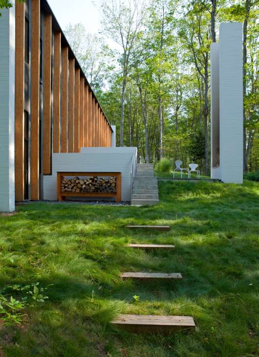 Modern house with white-painted walls and wooden slat screening nestled among tall forest trees, with a wooden stepping stone path leading toward the lake.
