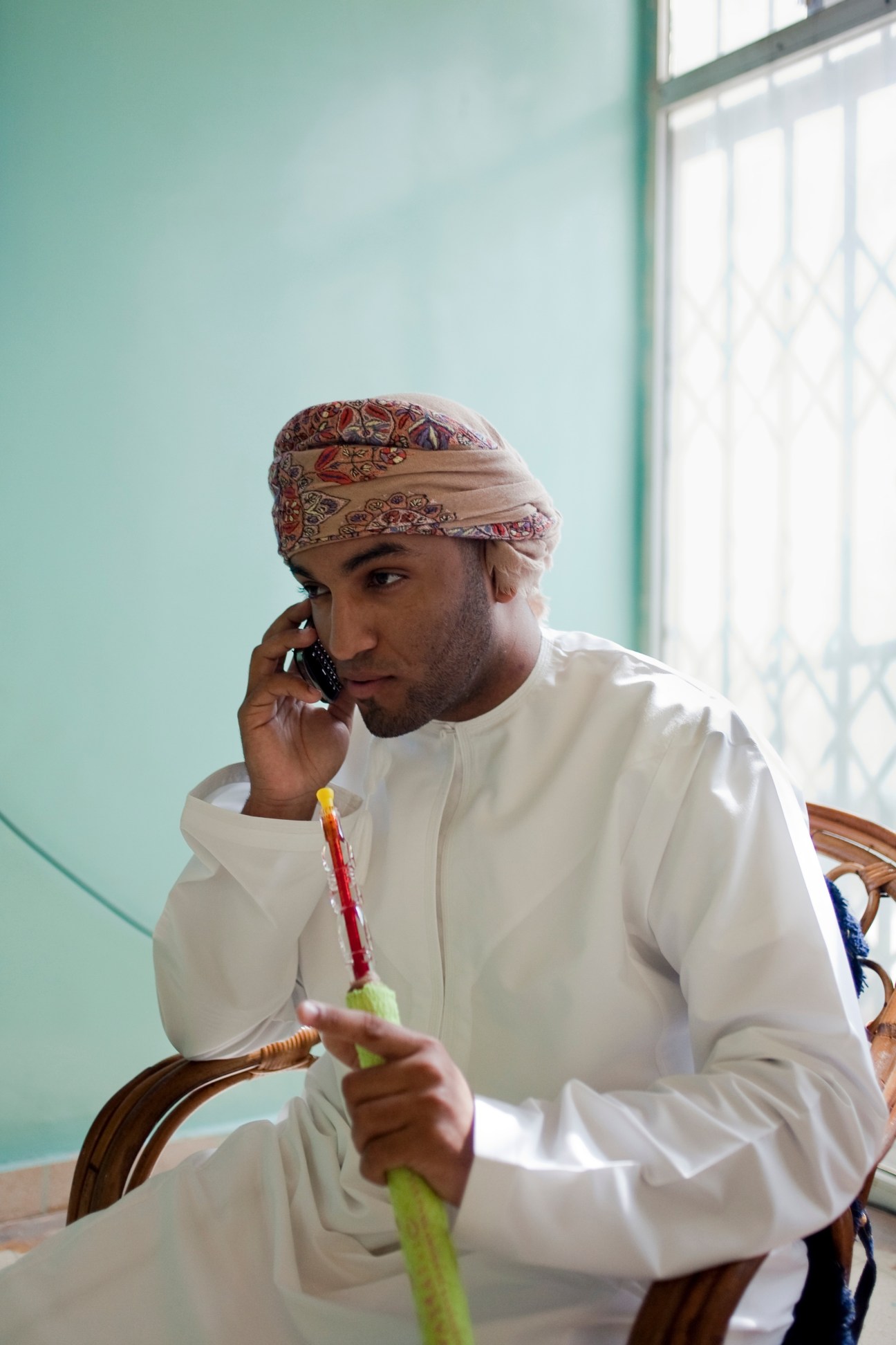 Man in traditional Omani dress speaking on phone while holding shisha pipe.
