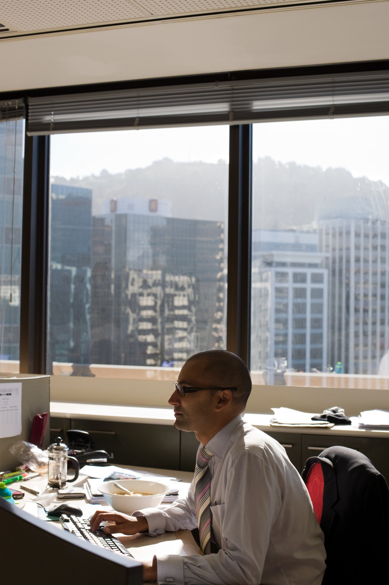 Man in business shirt at desk with cityscape view from high-rise office window.