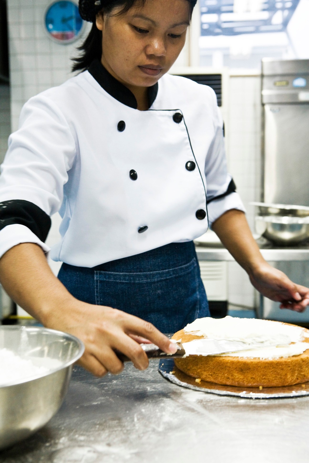 Filipino culinary student in white chef's coat preparing cake in training kitchen.