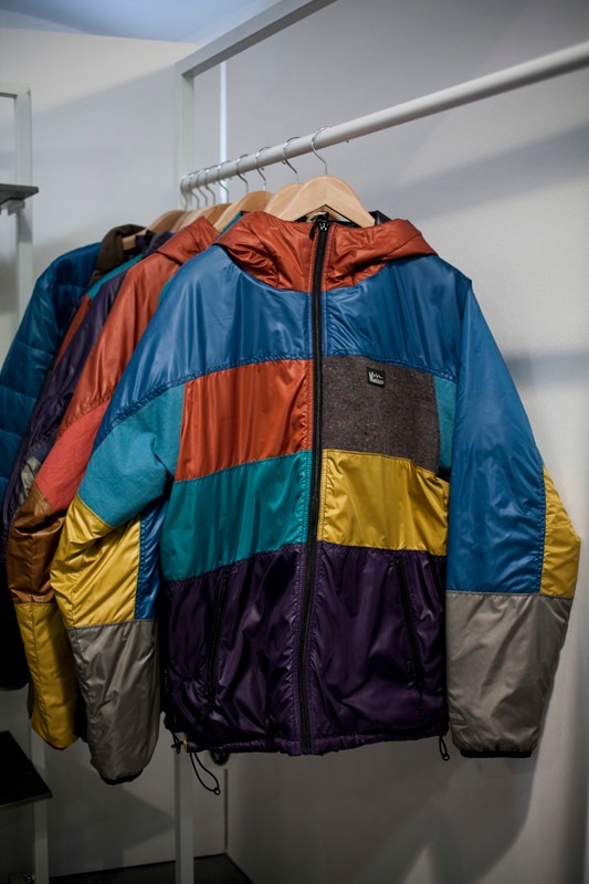 Manastash patchwork coat with colorful blocked sections in blue, orange, teal, yellow, and gray hanging on a rail.