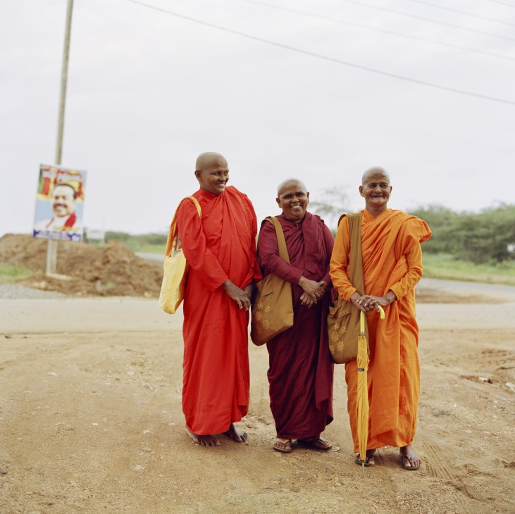Three female Buddhist monks in colorful robes walk together on a dirt road in Hambantota, Sri Lanka.