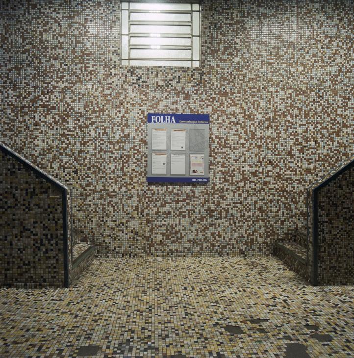 A hallway with small mosaic tiles in earth tones features a blue internal communications notice board labeled 'FOLHA'.